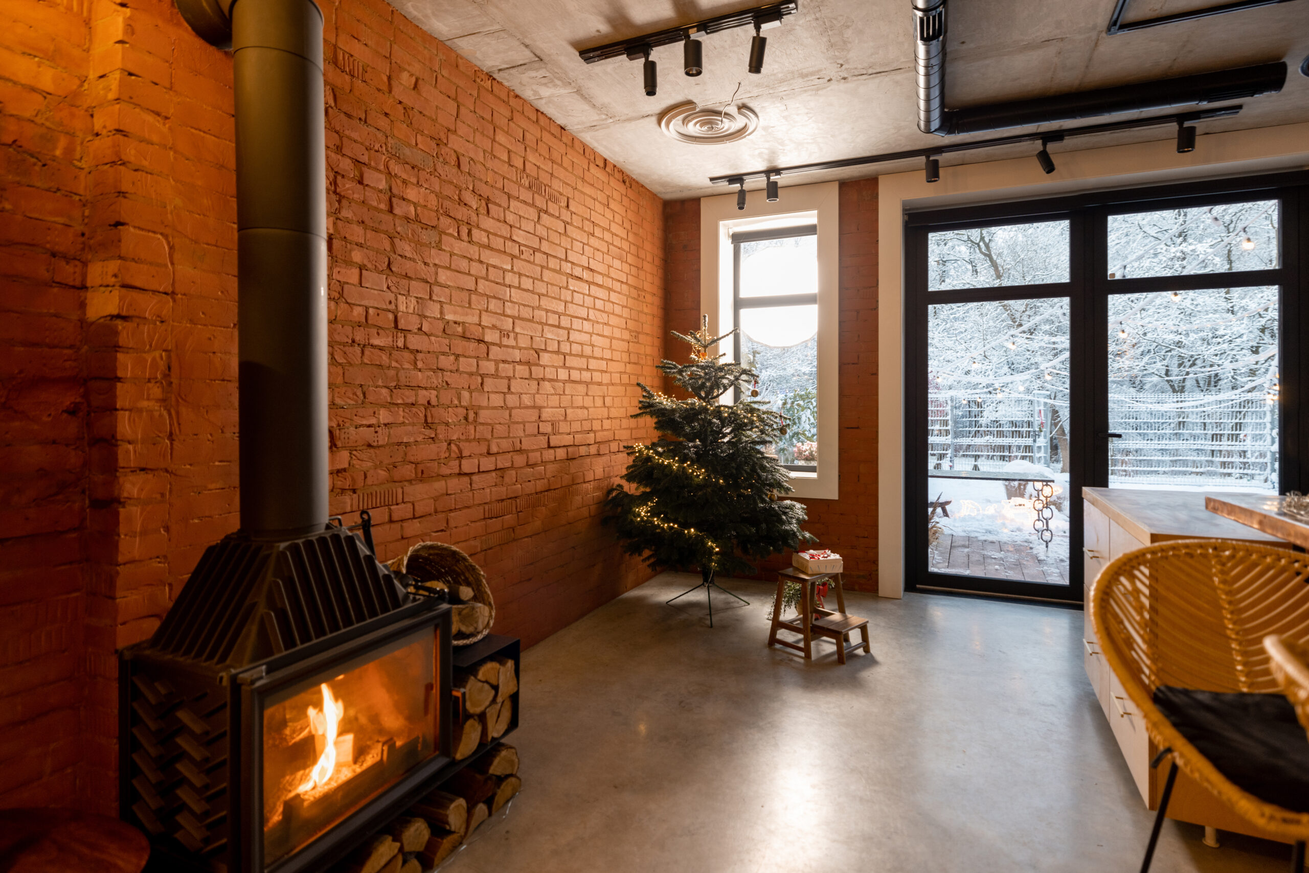 Cozy living room interior with Christmas tree and burning fireplace. Concept of home comfort in winter. Windows overlooking on snowy garden, room with brick wall and concrete floor